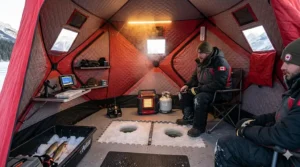 Interior view of a portable ice fishing tent featuring a heater, gear storage, and two anglers over drilled holes.